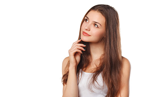 Junge Frau mit langen braunen Haaren, die ein weißes Tank-Top trägt und mit einem leichten Lächeln nachdenklich aufblickt, wobei sie mit einer Hand ihr Kinn berührt, und vor einem schlichten weißen Hintergrund steht.